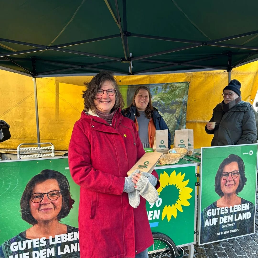 Eine Frau in rotem Mantel lächelt, während sie an einem Infostand mit dem Slogan ‚Gutes Leben auf dem Land‘ Flyer und Produkte präsentiert. Im Hintergrund stehen zwei weitere Personen unter einem Zelt.