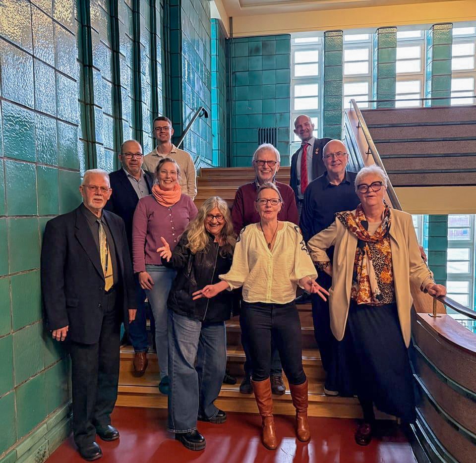 Gruppenfoto mit elf Personen auf einer Treppe vor grün gefliester Wand im historischen Gebäude. Mitglieder verschiedener politischer Gruppen aus Bremerhaven, darunter Vertreter*innen von Grünen, Die PARTEI, UWG, GfW und BASU.